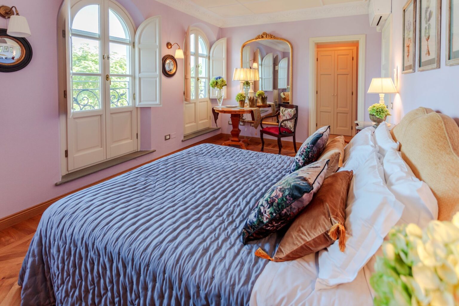 Cozy bedroom with a lavender wall, featuring a plush bed adorned with decorative pillows, large windows with white shutters, a wooden desk and chair, and a stylish mirror reflecting natural light. | Only on SopranoVillas
