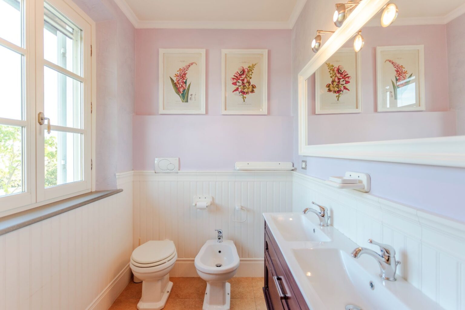 Bright and modern bathroom featuring a lavender wall color, double sink with elegant faucets, a toilet, and a bidet. The room is adorned with framed floral artwork and has large windows allowing natural light to enhance the space. | Only on SopranoVillas