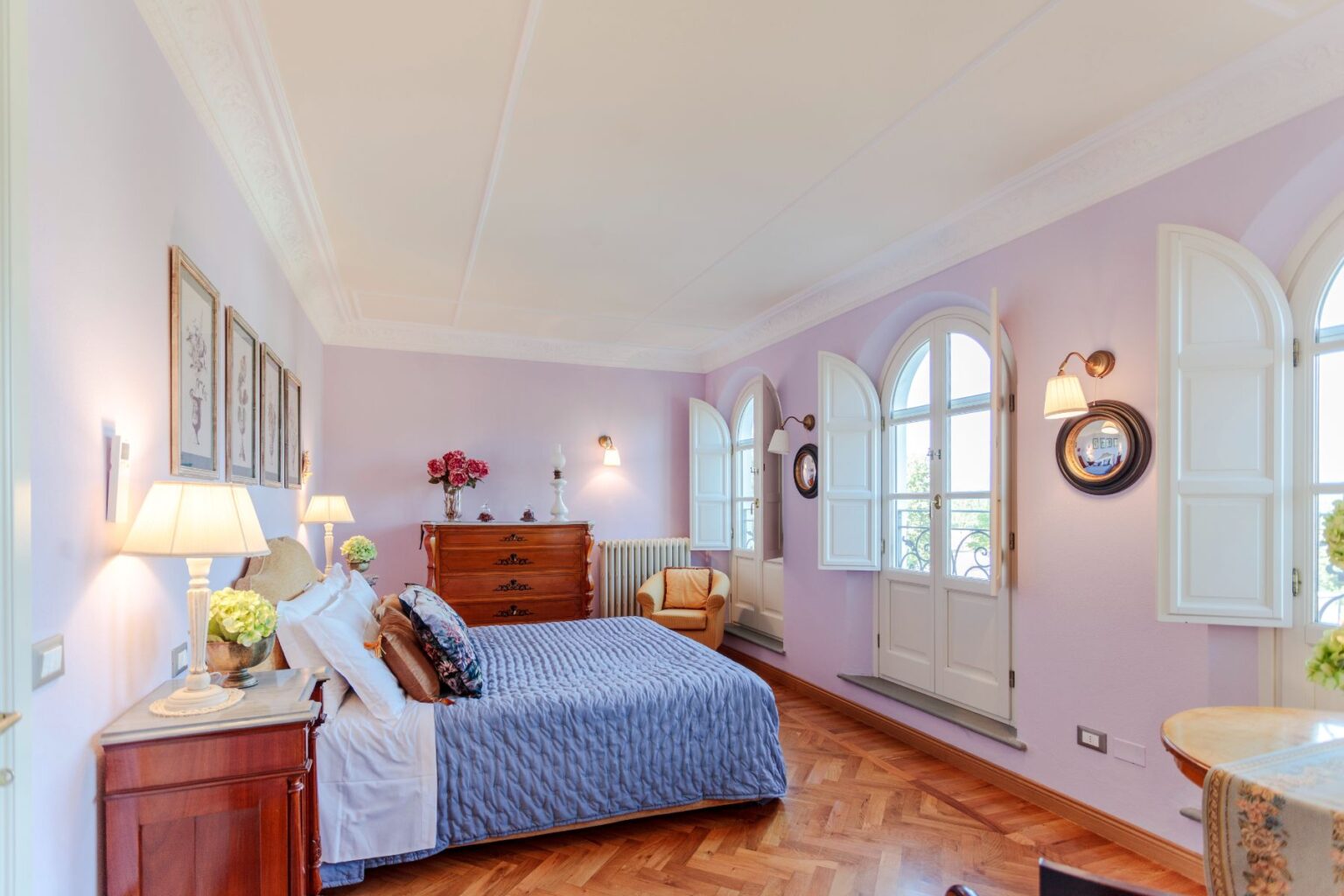 Cozy and elegantly designed bedroom featuring a queen-sized bed with a blue quilt, wooden furniture, and a pastel purple wall. The room includes large arched windows with white shutters, allowing natural light to brighten the space, and decorative elements like framed artwork and floral arrangements. Ideal for a relaxing getaway. | Only on SopranoVillas