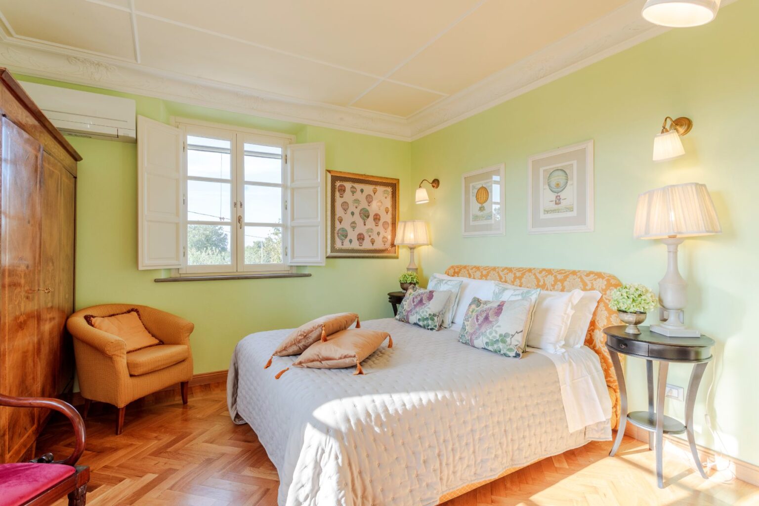 Cozy bedroom featuring a queen-sized bed with decorative pillows, soft green walls, and natural light from a window. The room includes a wooden wardrobe, a stylish armchair, and framed artwork, creating a warm and inviting atmosphere. | Only on SopranoVillas