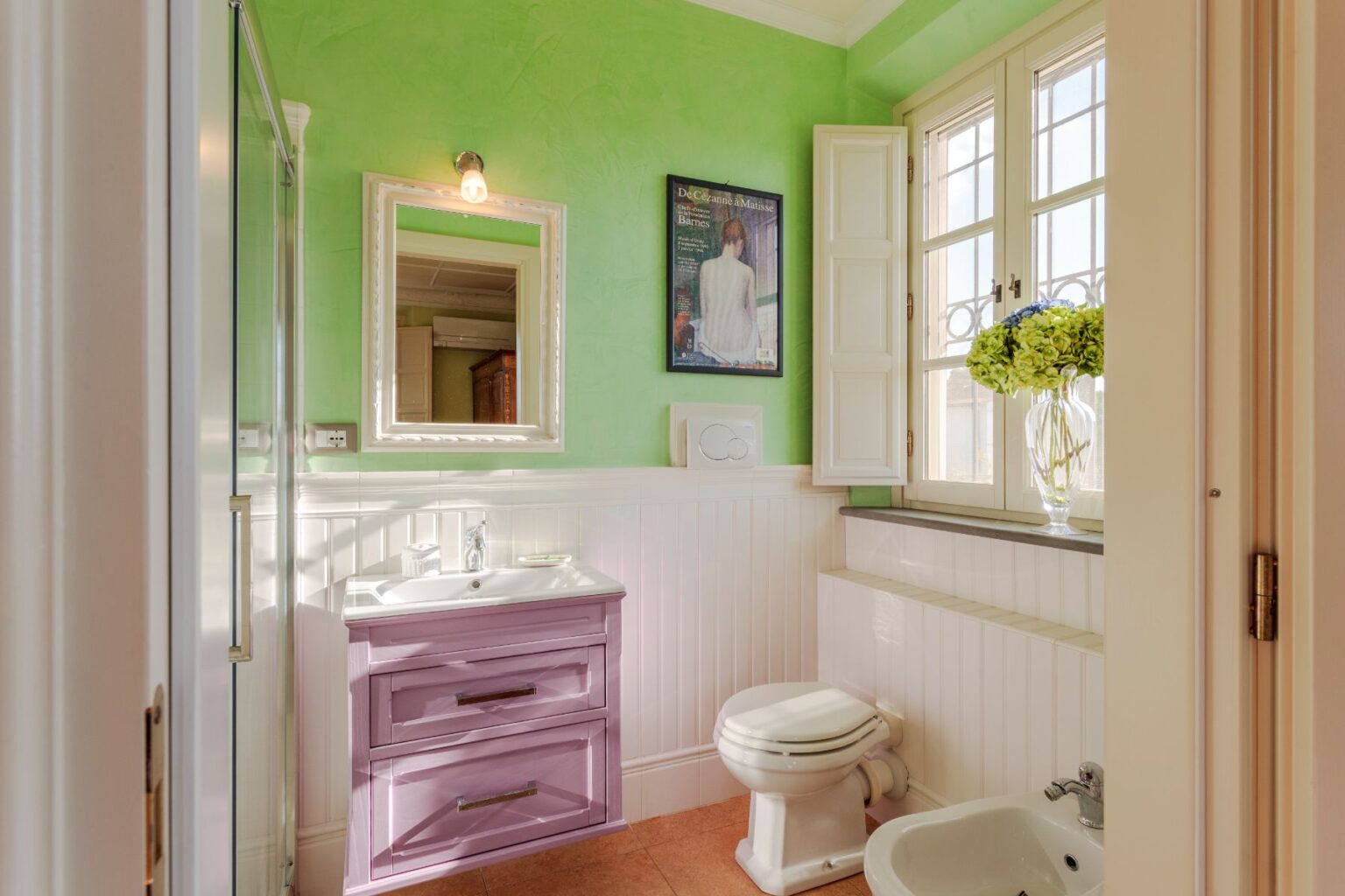 Bright and modern bathroom featuring a vibrant green wall, a stylish purple vanity with a sink, and a decorative mirror. Natural light streams in through a window adorned with white shutters, showcasing a floral arrangement. The space includes a toilet and bidet, complemented by elegant tile flooring. | Only on SopranoVillas