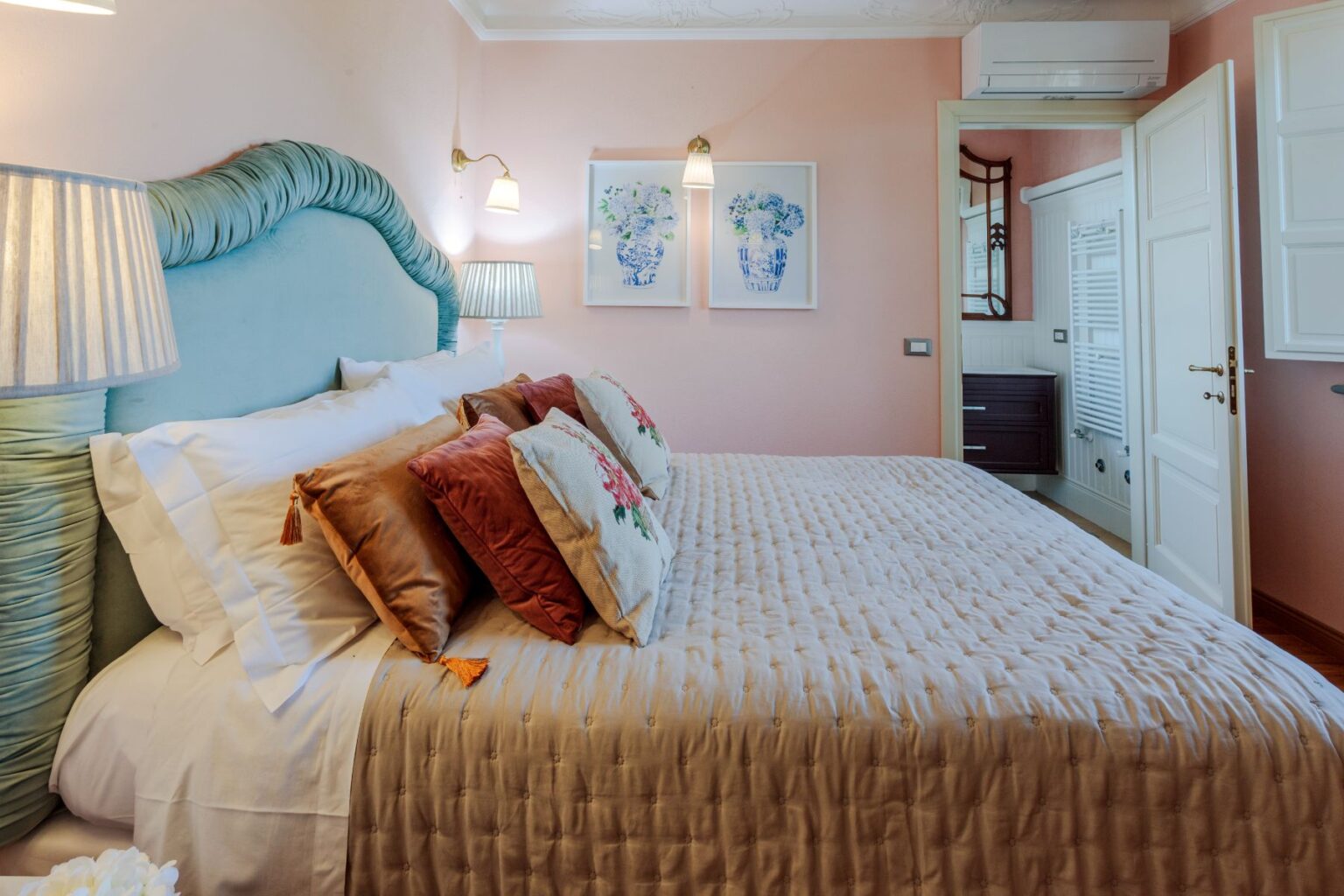 Cozy bedroom featuring a plush bed with decorative pillows, soft lighting from bedside lamps, and pastel pink walls. The room includes framed floral artwork and modern furnishings, creating a serene and inviting atmosphere. | Only on SopranoVillas