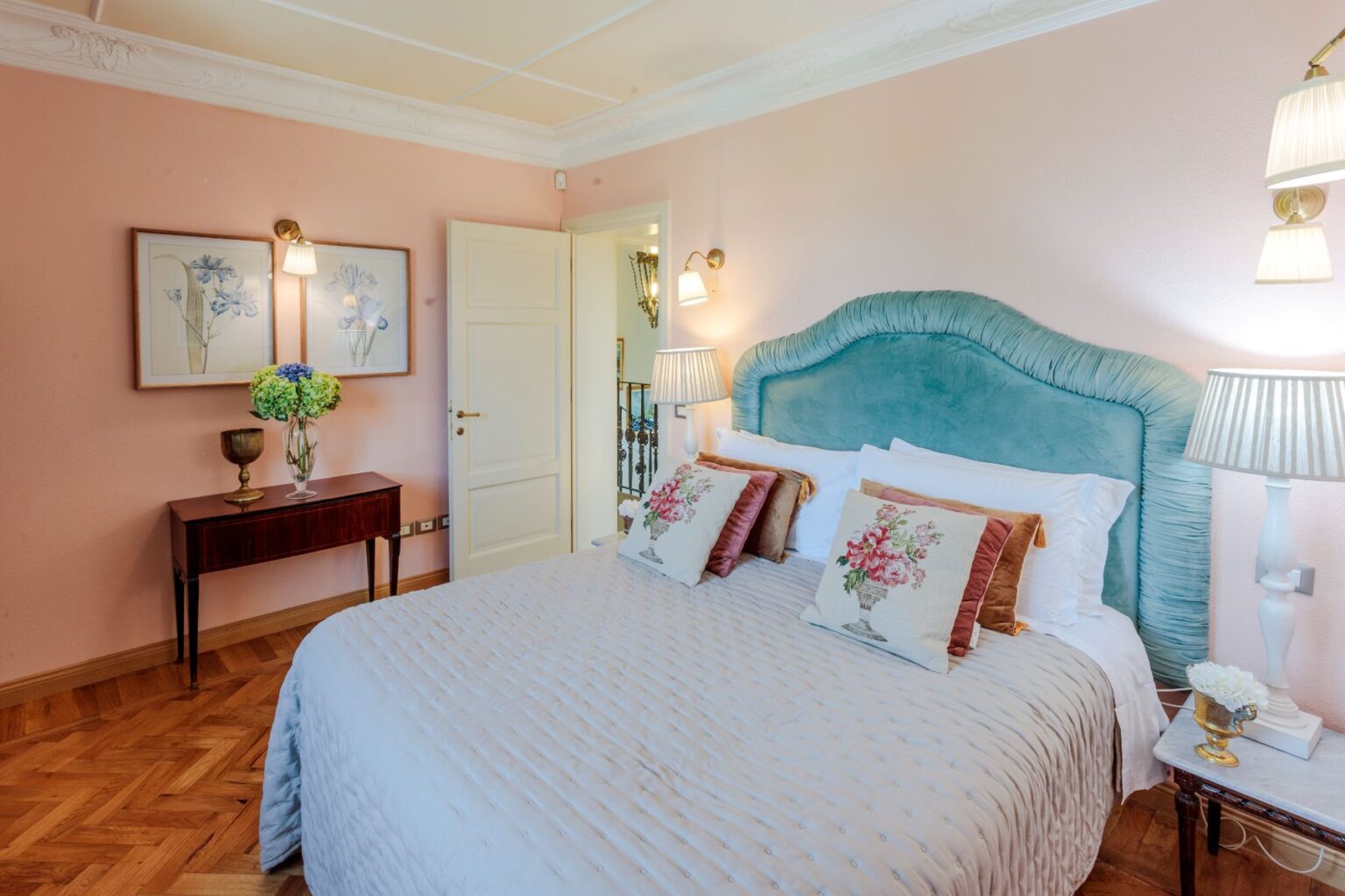 Elegant bedroom featuring a plush teal headboard, decorative pillows, and a cozy quilt. The room is adorned with floral artwork, a wooden bedside table, and stylish lamps, all set against soft pink walls and warm wooden flooring. | Only on SopranoVillas
