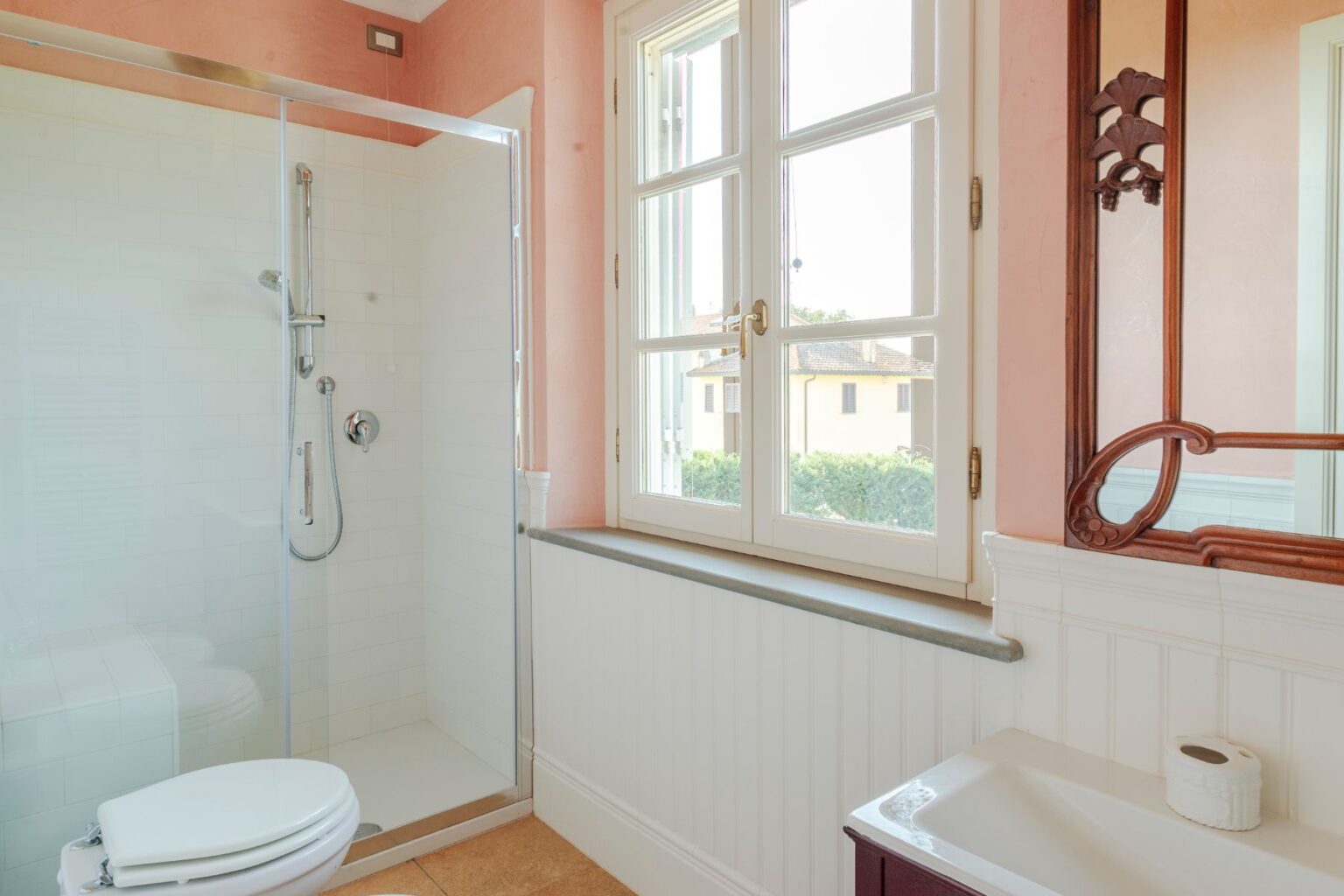 Modern bathroom featuring a glass shower enclosure, white tiles, and a window with a view. The space includes a toilet and a sink with a decorative mirror, accented by soft pink walls and a gray countertop. Ideal for showcasing contemporary bathroom design. | Only on SopranoVillas