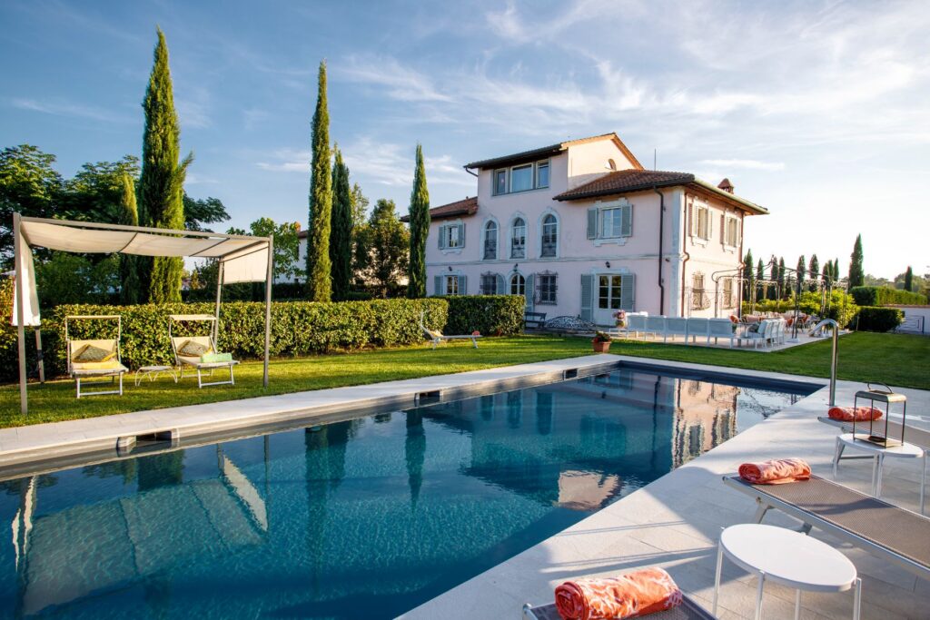 Luxurious villa featuring a serene swimming pool, sun loungers, and landscaped gardens, set against a clear blue sky. The elegant pink building showcases classic architectural details and is surrounded by lush greenery, creating a perfect outdoor relaxation space. | Only on SopranoVillas