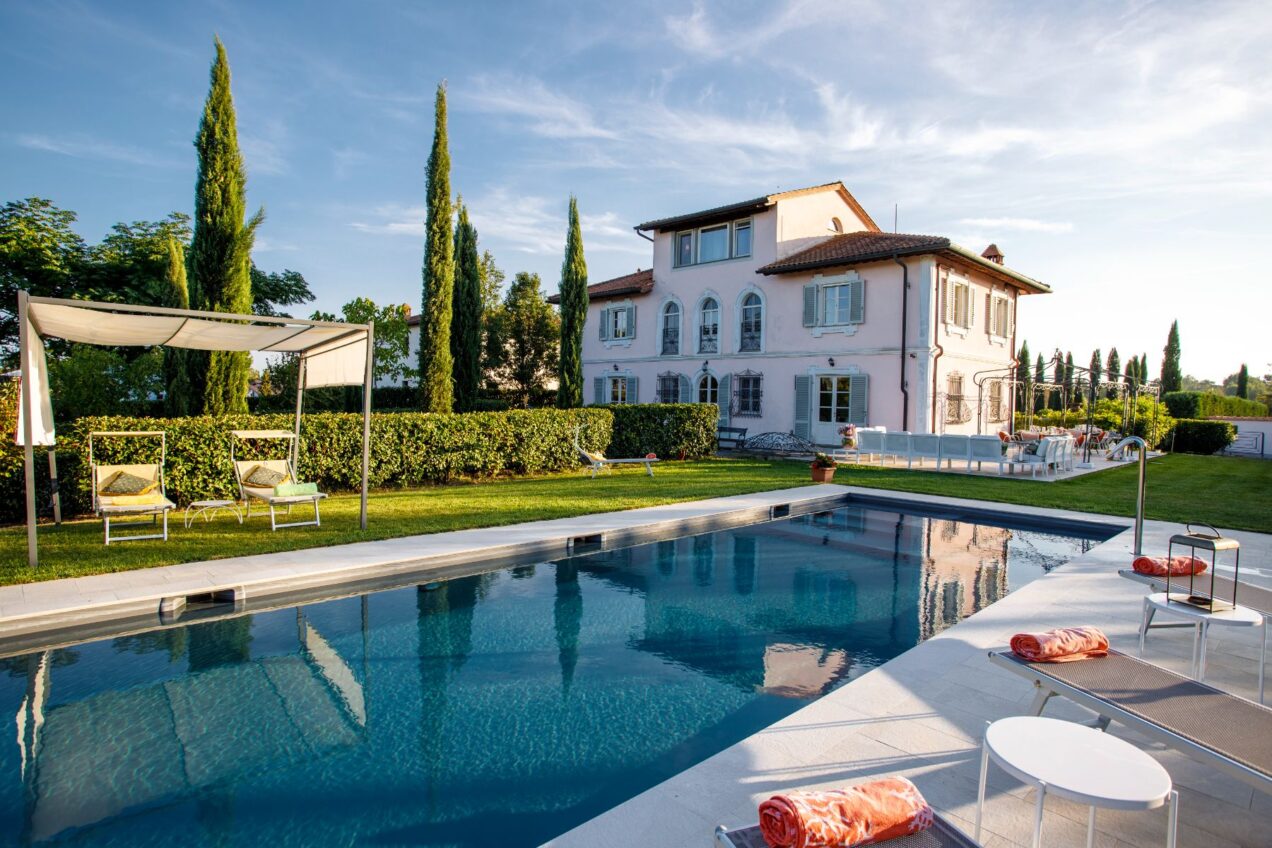 Luxurious villa featuring a serene swimming pool, sun loungers, and landscaped gardens, set against a clear blue sky. The elegant pink building showcases classic architectural details and is surrounded by lush greenery, creating a perfect outdoor relaxation space. | Only on SopranoVillas