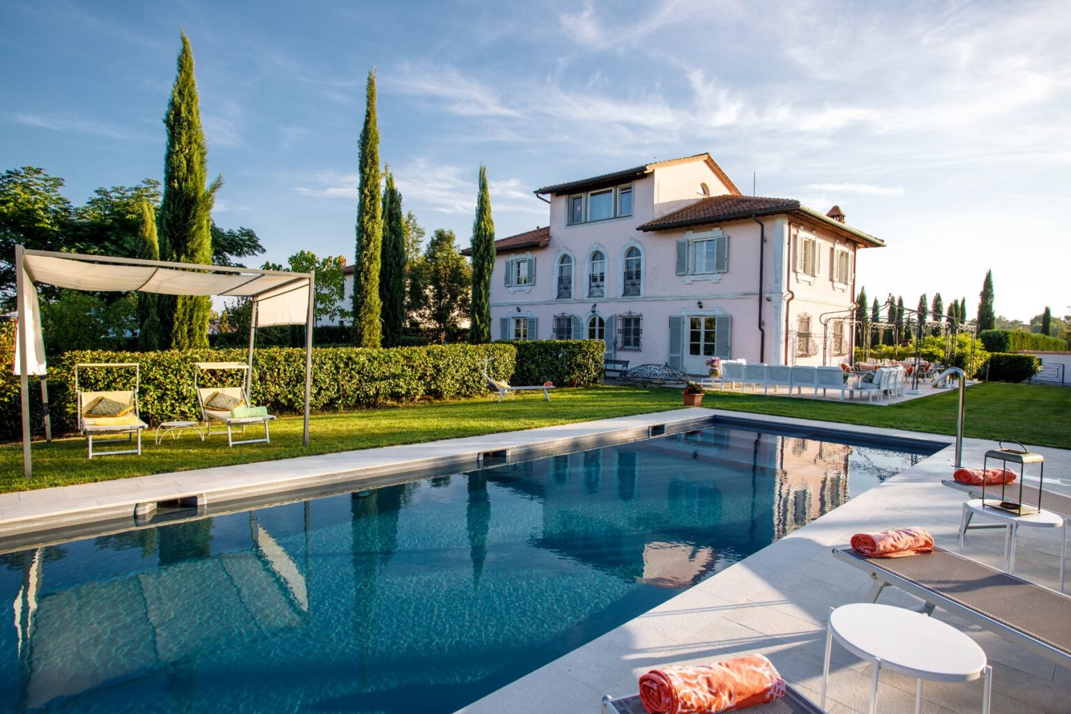 Luxurious villa featuring a serene swimming pool, sun loungers, and landscaped gardens, set against a clear blue sky. The elegant pink building showcases classic architectural details and is surrounded by lush greenery, creating a perfect outdoor relaxation space. | Only on SopranoVillas