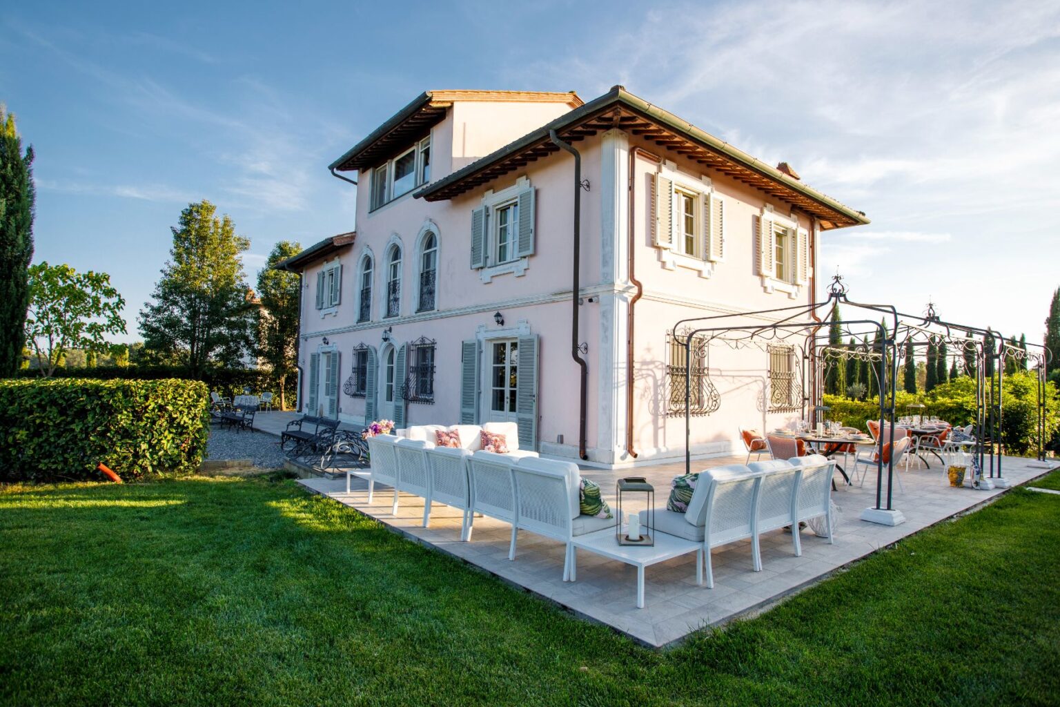 Elegant pink villa with lush green lawn, surrounded by trees, featuring a spacious patio with white seating and a dining area, perfect for outdoor gatherings. | Only on SopranoVillas