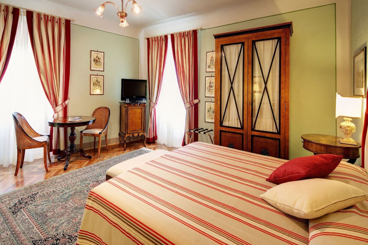 Cozy bedroom featuring a large striped bed, wooden wardrobe, and elegant decor. The room includes a small dining table with two chairs, a TV, and large windows draped with red and cream curtains, creating a warm, inviting atmosphere. | Only on SopranoVillas