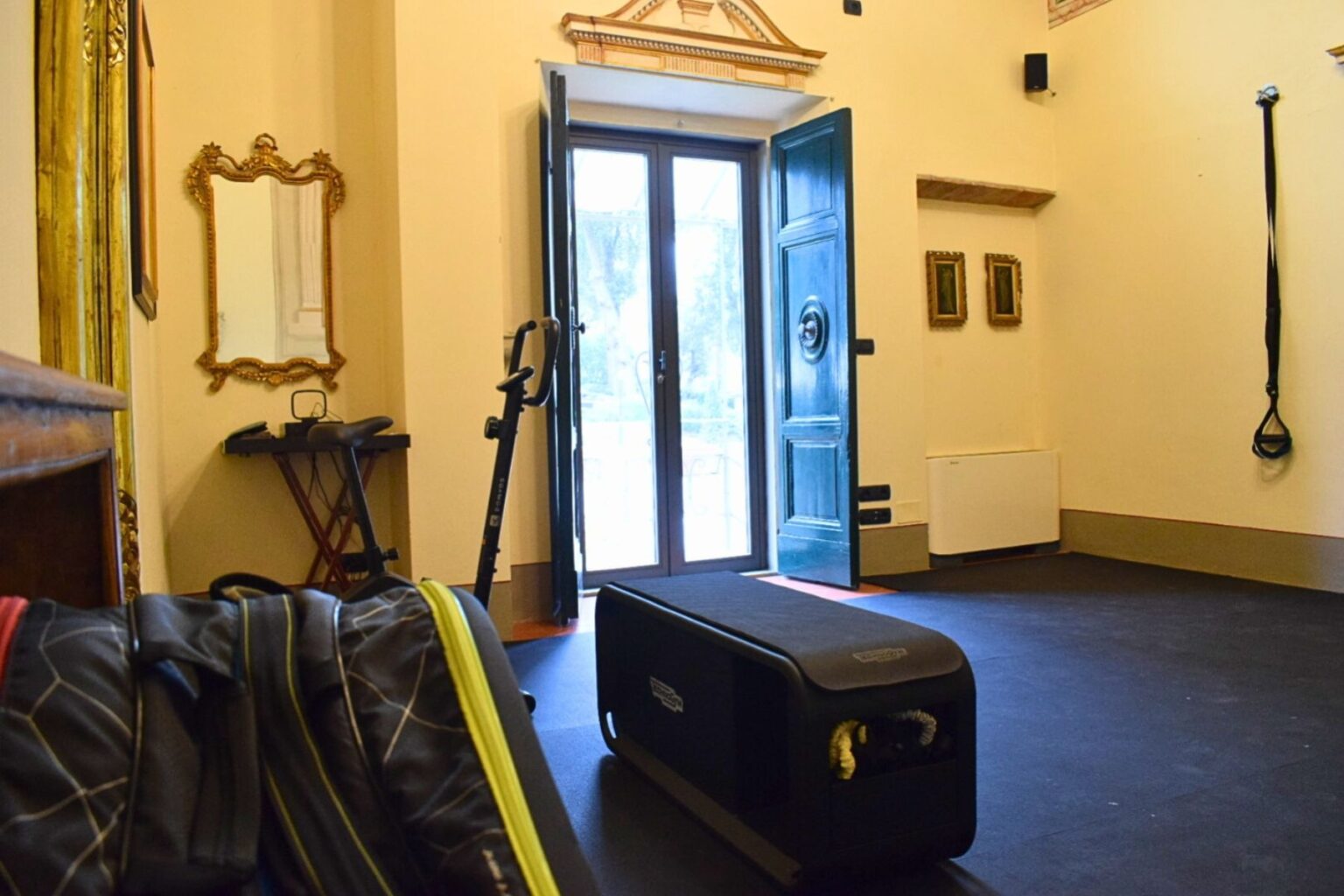 Home gym interior featuring an open door, exercise equipment, and workout gear. The room includes a mirrored wall, a small table, and fitness straps, creating a functional and inviting space for workouts. | Only on SopranoVillas