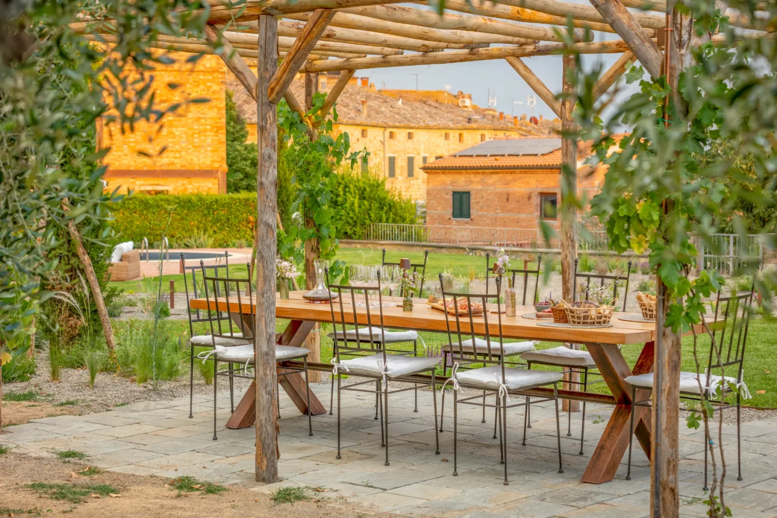 Outdoor dining area under a wooden pergola, featuring a long wooden table set with dishes and decorative elements, surrounded by lush greenery and rustic stone walls, ideal for gatherings and events. | Only on SopranoVillas