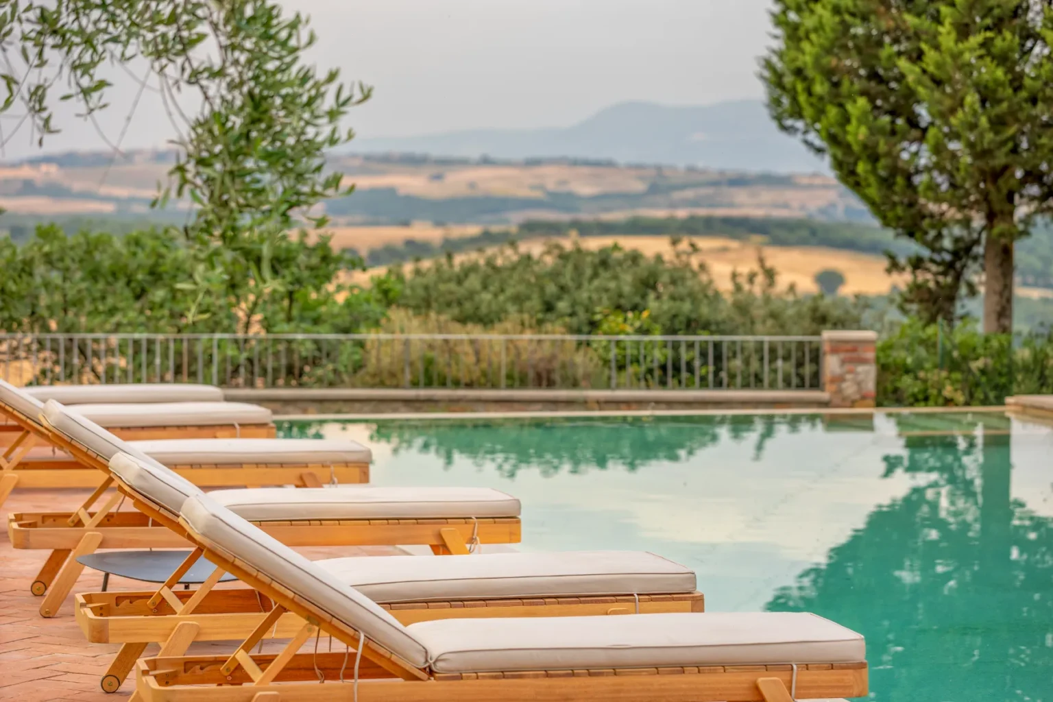 Relaxing outdoor pool area with wooden loungers and a scenic view of rolling hills and greenery, perfect for vacation or leisure activities. | Only on SopranoVillas