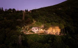 Aerial view of a modern villa nestled in a lush green hillside, featuring a swimming pool and outdoor seating areas, surrounded by trees and illuminated by soft evening lights.