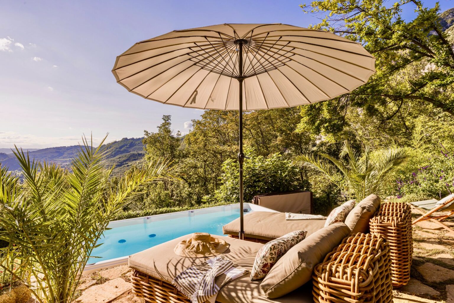 Luxurious outdoor lounge area with a large umbrella, comfortable cushions, and a stunning view of a swimming pool surrounded by lush greenery and mountains. Perfect for relaxation and enjoying nature. | Only on SopranoVillas