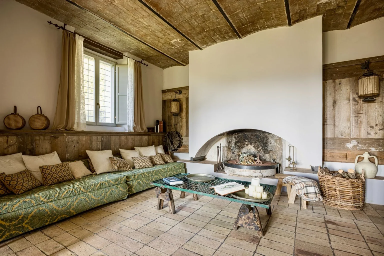 Cozy living room with a green patterned sofa adorned with decorative pillows, a rustic coffee table, and a traditional fireplace. Natural light streams through a window with sheer curtains, highlighting wooden accents and earthy tones in the decor. | Only on SopranoVillas