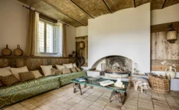 Cozy living room with a green patterned sofa adorned with decorative pillows, a rustic coffee table, and a traditional fireplace. Natural light streams through a window with sheer curtains, highlighting wooden accents and earthy tones in the decor.