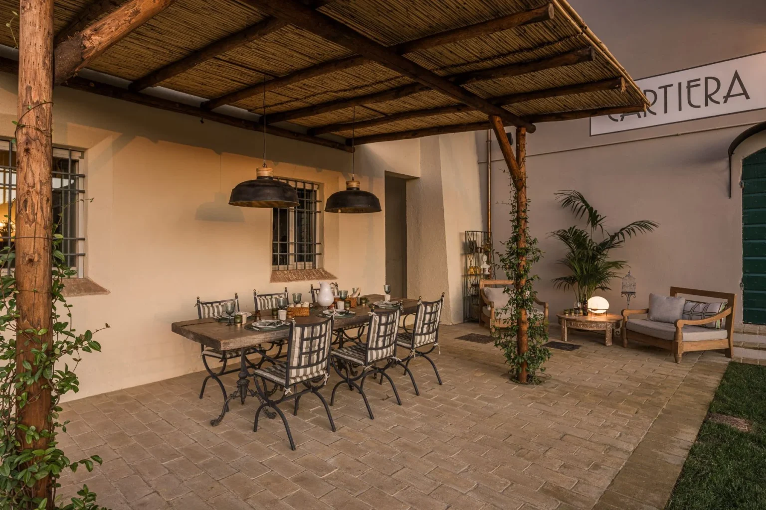 Outdoor dining area featuring a rustic wooden table with metal chairs, set under a thatched roof. The space is adorned with hanging pendant lights, potted plants, and decorative elements, creating a cozy ambiance for gatherings. | Only on SopranoVillas