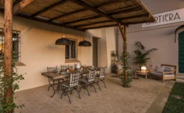 Outdoor dining area featuring a rustic wooden table with metal chairs, set under a thatched roof. The space is adorned with hanging pendant lights, potted plants, and decorative elements, creating a cozy ambiance for gatherings.