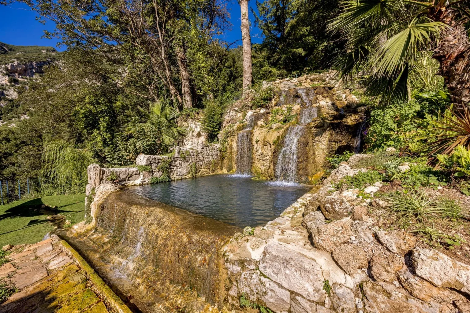 Natural stone waterfall cascading into a serene pool surrounded by lush greenery and palm trees, set against a clear blue sky. Ideal for relaxation and nature photography. | Only on SopranoVillas