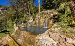 Natural stone waterfall cascading into a serene pool surrounded by lush greenery and palm trees, set against a clear blue sky. Ideal for relaxation and nature photography.