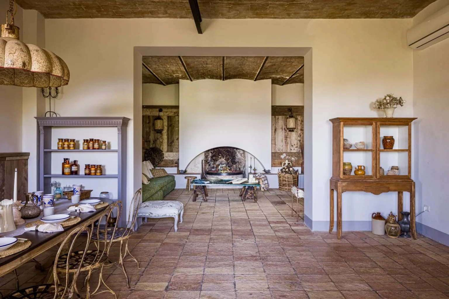 Interior view of a stylish living space featuring a rustic wooden dining table set for a meal, vintage chairs, and a cozy green sofa. The room includes a large fireplace, decorative shelves filled with jars, and warm lighting, showcasing a blend of modern and traditional design elements. | Only on SopranoVillas