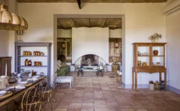 Interior view of a stylish living space featuring a rustic wooden dining table set for a meal, vintage chairs, and a cozy green sofa. The room includes a large fireplace, decorative shelves filled with jars, and warm lighting, showcasing a blend of modern and traditional design elements.
