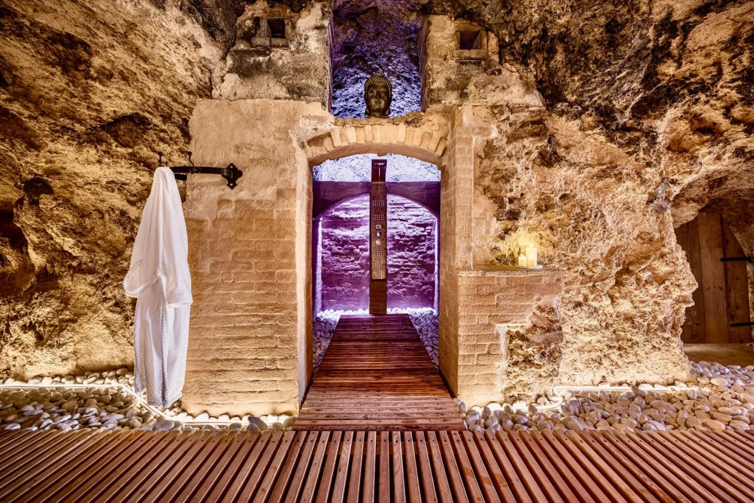 Serene spa entrance in a cave-like setting with natural stone walls, wooden flooring, and soft ambient lighting, featuring a white robe hanging on a hook and candles placed on a stone ledge. | Only on SopranoVillas