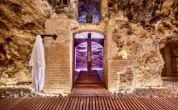 Serene spa entrance in a cave-like setting with natural stone walls, wooden flooring, and soft ambient lighting, featuring a white robe hanging on a hook and candles placed on a stone ledge.