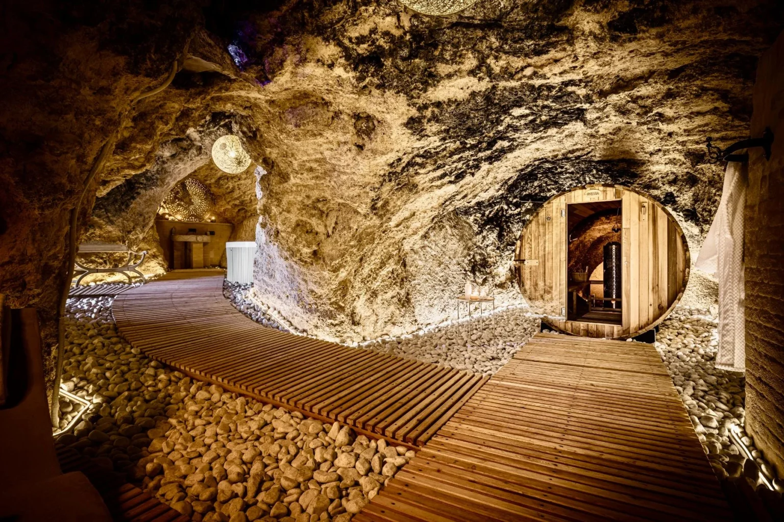 Interior view of a unique cave spa featuring wooden pathways, smooth stones, and soft lighting, with a circular wooden sauna and a cozy relaxation area, showcasing natural rock formations and an inviting atmosphere for wellness and relaxation. | Only on SopranoVillas