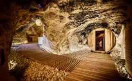 Interior view of a unique cave spa featuring wooden pathways, smooth stones, and soft lighting, with a circular wooden sauna and a cozy relaxation area, showcasing natural rock formations and an inviting atmosphere for wellness and relaxation.