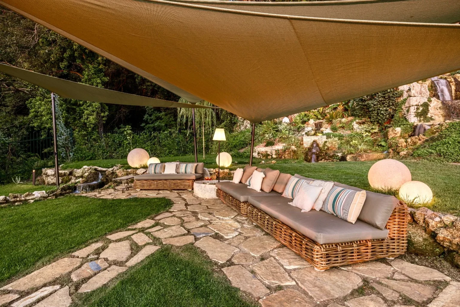 Cozy outdoor seating area featuring wicker sofas with cushions under a large shade sail, surrounded by lush greenery and a stone pathway, creating a serene garden retreat. | Only on SopranoVillas