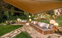 Cozy outdoor seating area featuring wicker sofas with cushions under a large shade sail, surrounded by lush greenery and a stone pathway, creating a serene garden retreat.