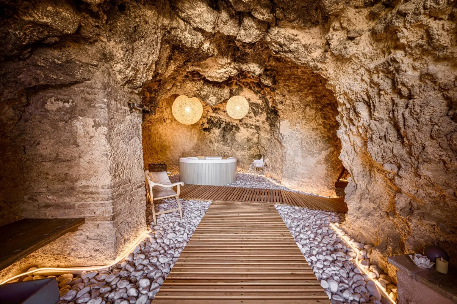 Relaxing spa interior featuring a natural cave setting, wooden walkway, smooth pebbles, and a modern hot tub, illuminated by stylish pendant lights. Ideal for wellness and tranquility. | Only on SopranoVillas