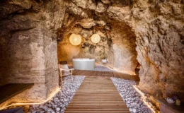 Relaxing spa interior featuring a natural cave setting, wooden walkway, smooth pebbles, and a modern hot tub, illuminated by stylish pendant lights. Ideal for wellness and tranquility.