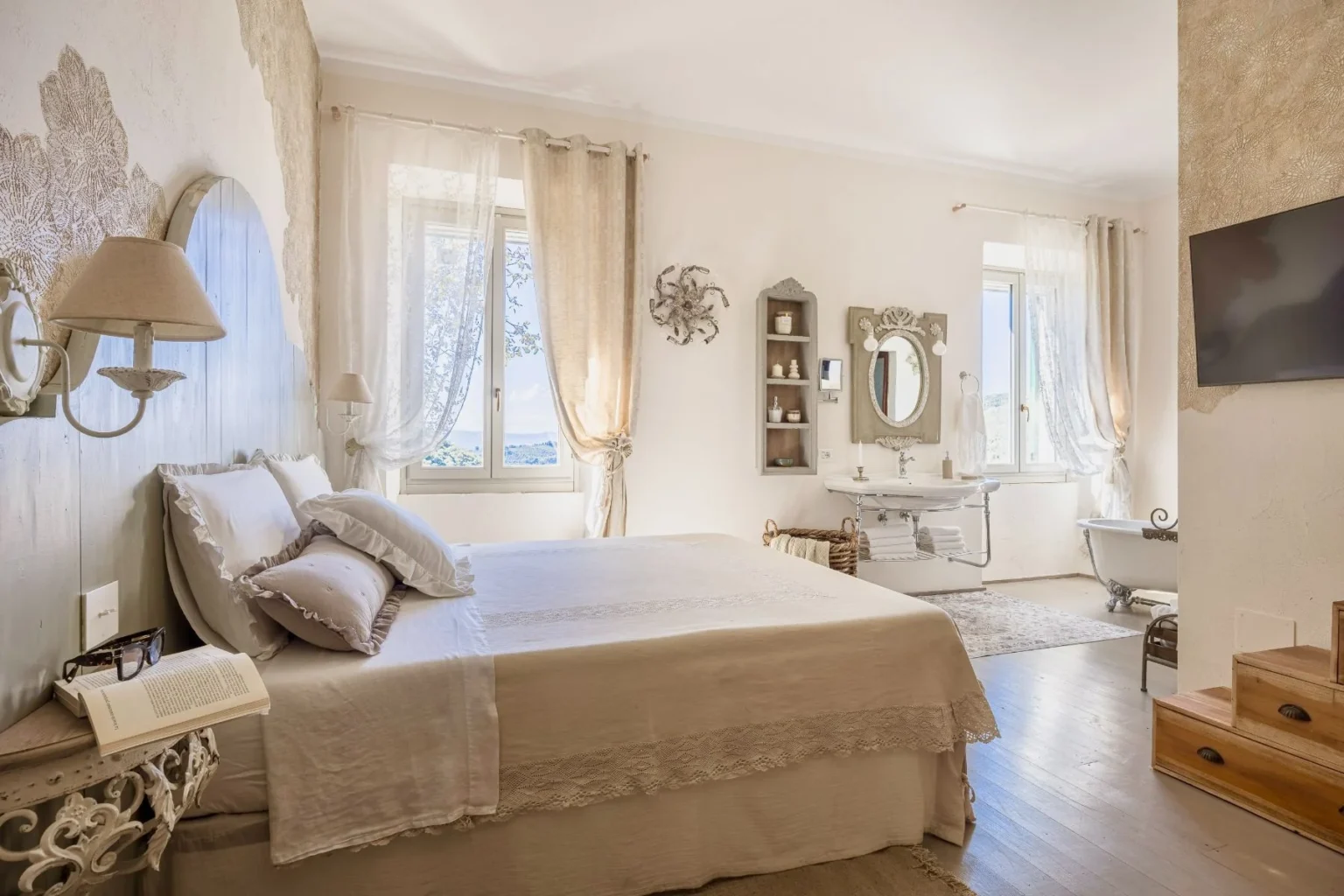 Bright and airy bedroom featuring a large bed with decorative pillows, elegant wall decor, and natural light streaming through sheer curtains. The room includes a vintage-style wash basin, a clawfoot bathtub, and tasteful furnishings, creating a serene and inviting atmosphere. Ideal for relaxation and comfort. | Only on SopranoVillas