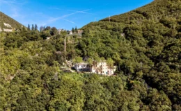 Aerial view of a secluded villa surrounded by lush greenery on a hillside, with clear blue skies and distant homes visible in the background.