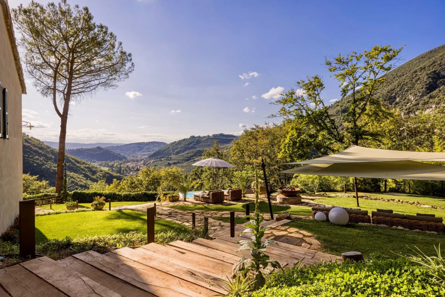 Scenic view of a landscaped garden with a wooden pathway leading to a cozy seating area, surrounded by lush greenery and mountains under a clear blue sky, featuring an umbrella and decorative stones. Ideal for outdoor relaxation and nature appreciation. | Only on SopranoVillas