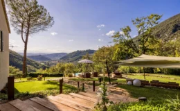 Scenic view of a landscaped garden with a wooden pathway leading to a cozy seating area, surrounded by lush greenery and mountains under a clear blue sky, featuring an umbrella and decorative stones. Ideal for outdoor relaxation and nature appreciation.