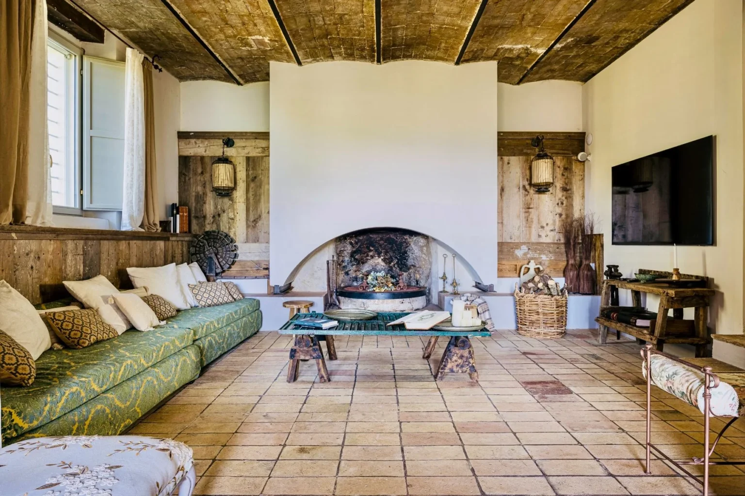 Cozy living room featuring a green patterned sofa with decorative pillows, a rustic wooden ceiling, and a traditional fireplace. The space includes a wooden coffee table, a basket of logs, and a modern TV, creating a blend of rustic charm and contemporary comfort. Natural light streams in from a window, enhancing the inviting atmosphere. | Only on SopranoVillas