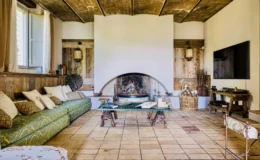Cozy living room featuring a green patterned sofa with decorative pillows, a rustic wooden ceiling, and a traditional fireplace. The space includes a wooden coffee table, a basket of logs, and a modern TV, creating a blend of rustic charm and contemporary comfort. Natural light streams in from a window, enhancing the inviting atmosphere.