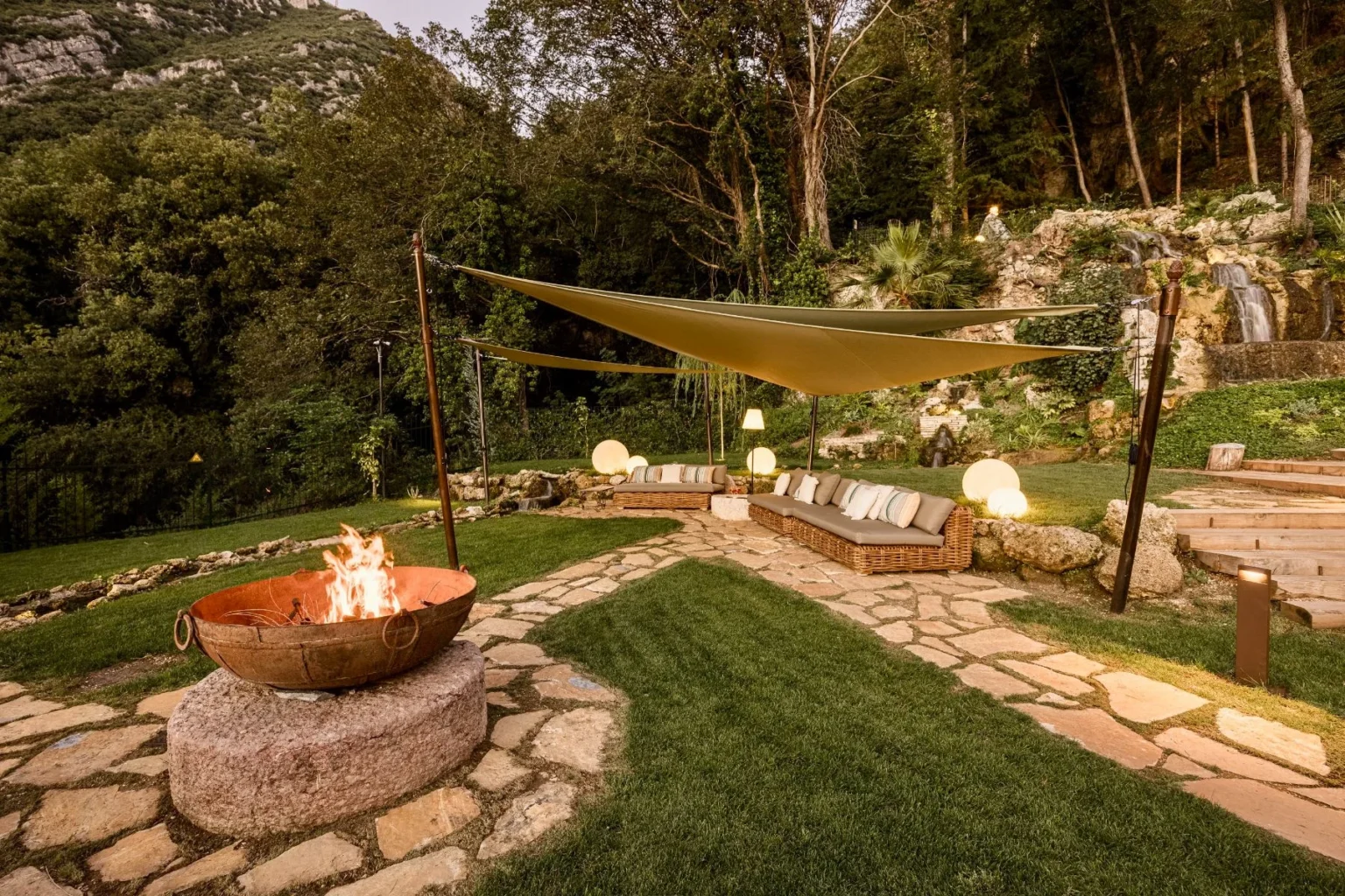 Cozy outdoor lounge area featuring a fire pit, comfortable seating under shade sails, and lush greenery in a serene garden setting. Ideal for relaxation and gatherings. | Only on SopranoVillas