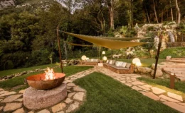 Cozy outdoor lounge area featuring a fire pit, comfortable seating under shade sails, and lush greenery in a serene garden setting. Ideal for relaxation and gatherings.