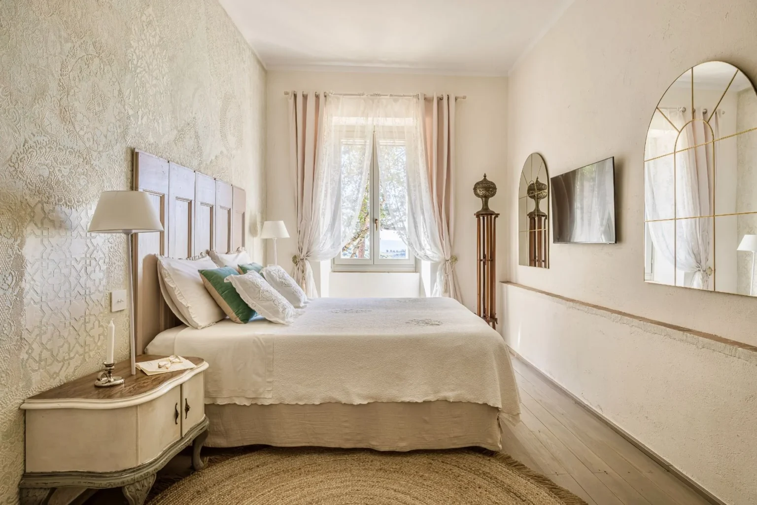 Cozy bedroom featuring a stylish wooden headboard, soft white bedding, and decorative pillows. Natural light floods the space through sheer curtains, highlighting the textured wallpaper and elegant mirrors. A bedside table with a lamp and a decorative rug complete the inviting atmosphere. | Only on SopranoVillas
