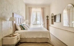 Cozy bedroom featuring a stylish wooden headboard, soft white bedding, and decorative pillows. Natural light floods the space through sheer curtains, highlighting the textured wallpaper and elegant mirrors. A bedside table with a lamp and a decorative rug complete the inviting atmosphere.