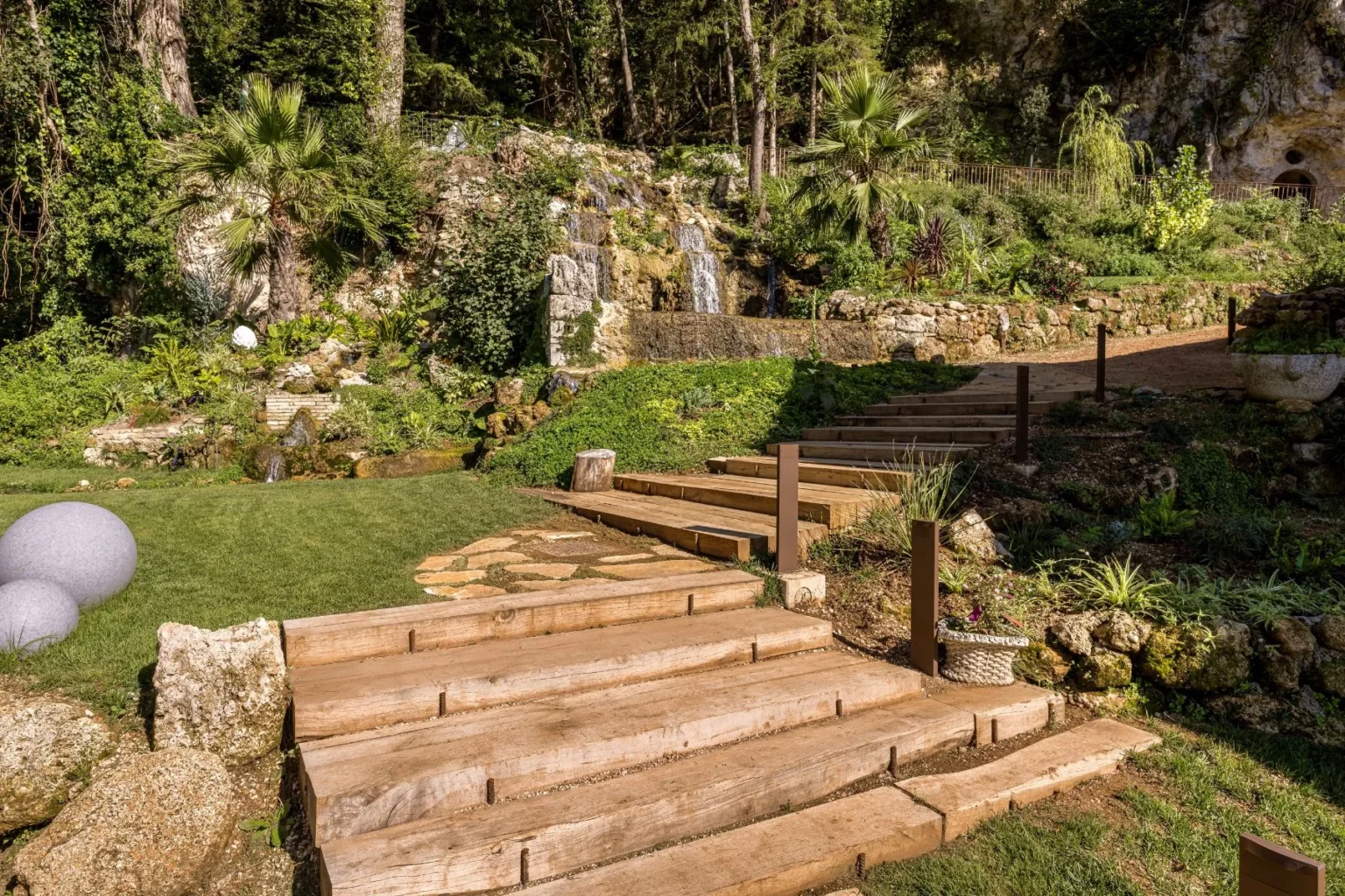 Lush garden landscape featuring a stone waterfall, wooden steps leading through vibrant greenery, and decorative round stones on a grassy area, ideal for outdoor relaxation and landscaping inspiration. | Only on SopranoVillas