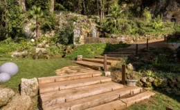 Lush garden landscape featuring a stone waterfall, wooden steps leading through vibrant greenery, and decorative round stones on a grassy area, ideal for outdoor relaxation and landscaping inspiration.
