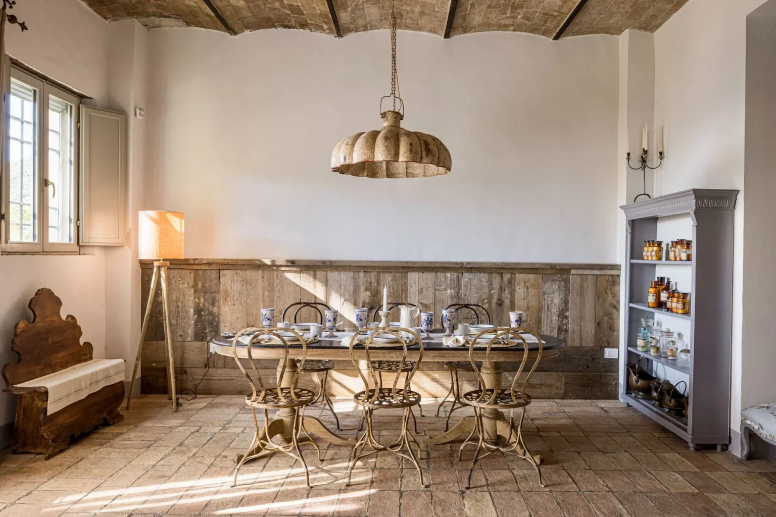 Stylish rustic dining room featuring a wooden table set for a meal, surrounded by vintage metal chairs. Natural light streams in through a window, illuminating a wooden sideboard filled with jars and a cozy floor lamp adds warmth to the space. | Only on SopranoVillas