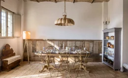 Stylish rustic dining room featuring a wooden table set for a meal, surrounded by vintage metal chairs. Natural light streams in through a window, illuminating a wooden sideboard filled with jars and a cozy floor lamp adds warmth to the space.