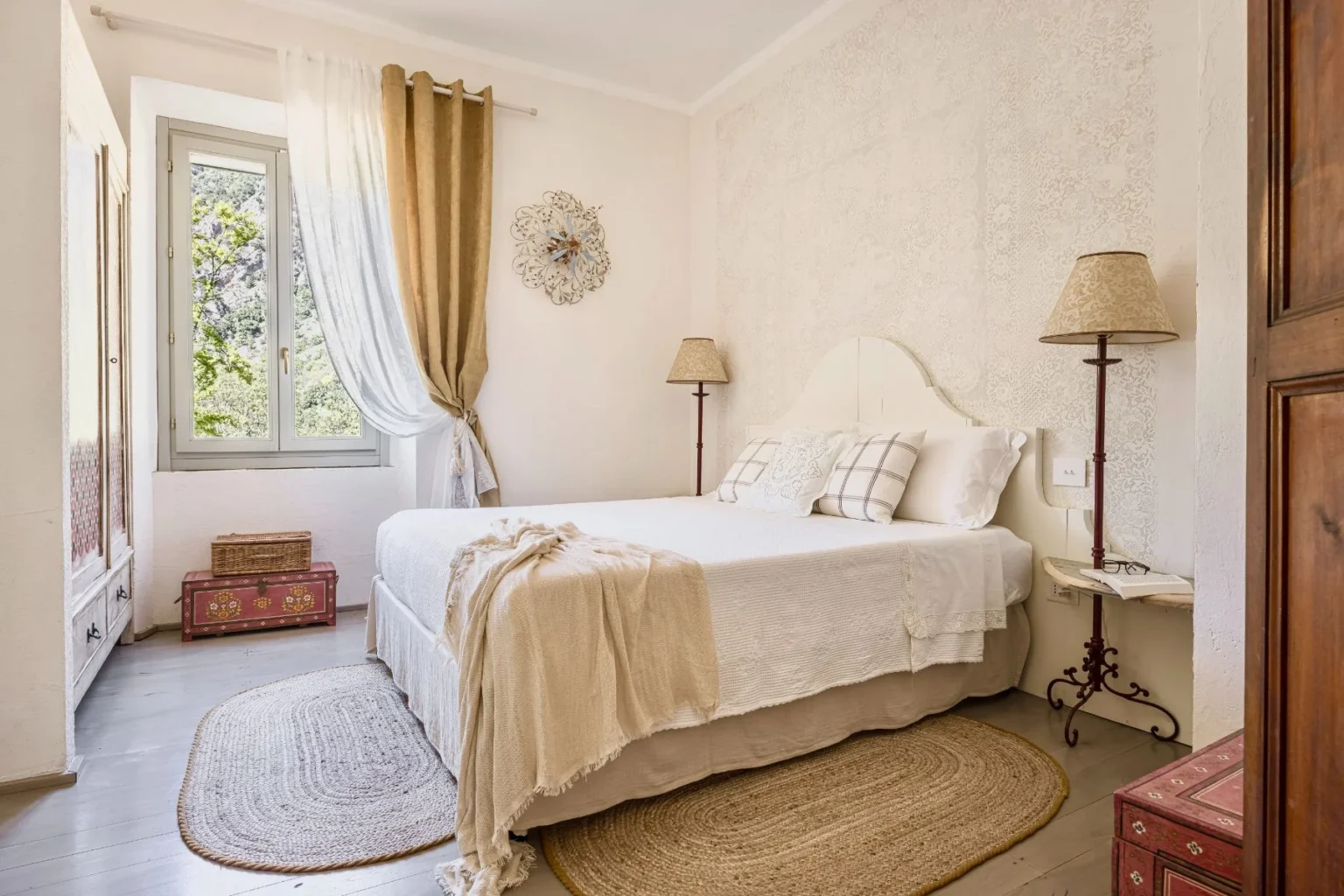 Cozy bedroom with a white bed adorned with decorative pillows, a vintage lamp, and a round woven rug, featuring natural light from a window dressed in elegant curtains. A wooden cabinet and colorful storage boxes add charm to the serene atmosphere. | Only on SopranoVillas