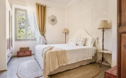Cozy bedroom with a white bed adorned with decorative pillows, a vintage lamp, and a round woven rug, featuring natural light from a window dressed in elegant curtains. A wooden cabinet and colorful storage boxes add charm to the serene atmosphere.